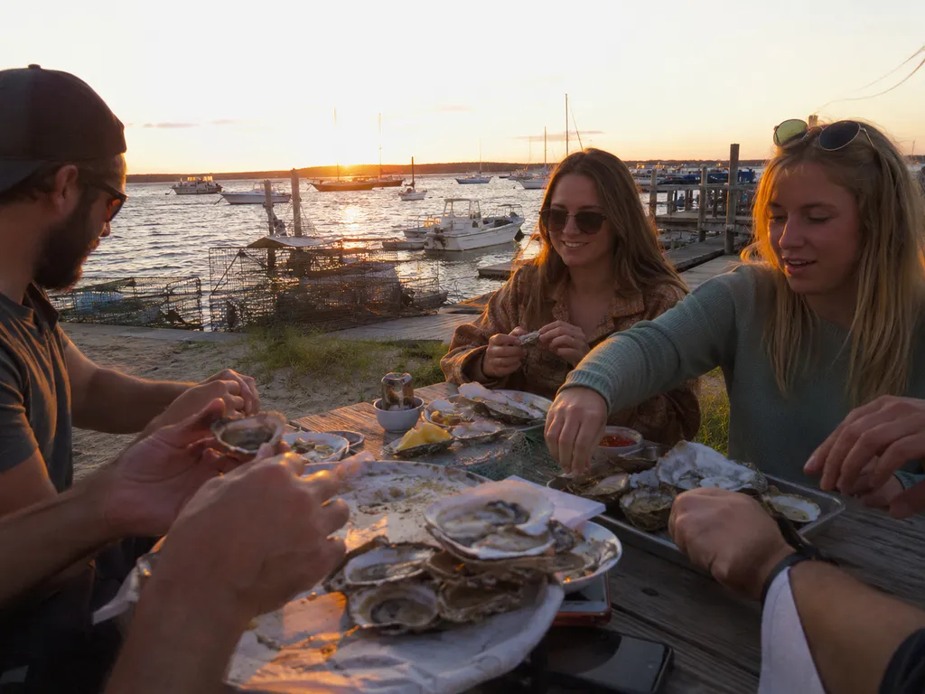 People eat oysters at a table by the water at sunset.